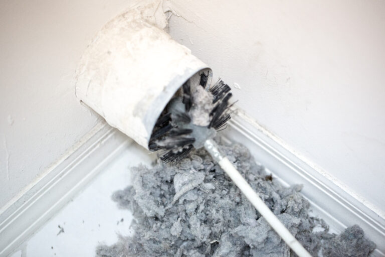 Dryer vent in a home being cleaned out with a round brush. There is a large pile of lint that has been removed from the vent on a white tiled floor. The walls and baseboards are white. The lint is gray. Taken with a Canon 5D Mark 3 camera.  rm